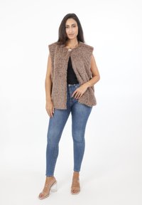 Kamizelka z futra syntetycznego w jasnobrązowym kolorze, bez kołnierza, z zapięciem z przodu. Połączona z niebieskimi, dopasowanymi dżinsami i przezroczystymi sandałami na obcasie.