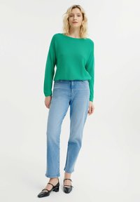 Grünes, texturiertes Langarmoberteil kombiniert mit hellblauen, geraden Jeans und schwarzen Absatzschuhen, die ein lässiges Outfit zeigen.