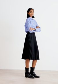 Blauwe en witte gestreepte blouse met lange mouwen, zwarte plooiskirt en zwarte enkelboots, tegen een effen witte achtergrond.