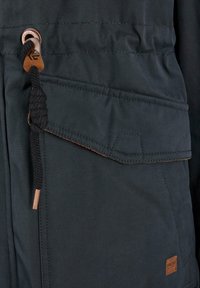 Veste bleu foncé avec une finition texturée, dotée d'une poche poitrine, de détails à cordon de serrage et d'un accent en cuir près de l'ourlet.