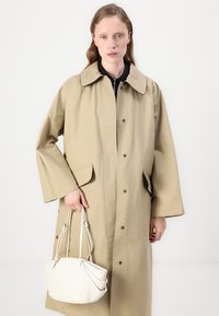 Beżowy bawełniany trench z dużym kołnierzem, bocznymi kieszeniami i napami; trzymany z małą, białą plisowaną torebką.