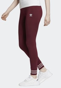 Bordowe legginsy wykonane z elastycznego materiału, z białym logo Adidas i białymi paskami na kostce. Gładka tekstura.