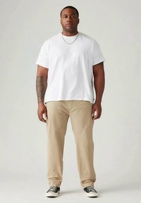 White short-sleeve t-shirt, beige pants, black and white shoes. Tattoo on left arm, standing against a neutral background.