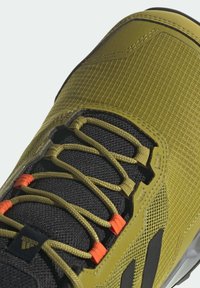 Gros plan d'une basket vert olive et noire avec tissu texturé, lacets olives, accents orange et logo Adidas sur la languette.