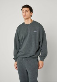 Sweatshirt gris foncé et confortable avec un col rond et des épaules tombantes, présentant un petit patch logo sur la poitrine et des poignets côtelés.