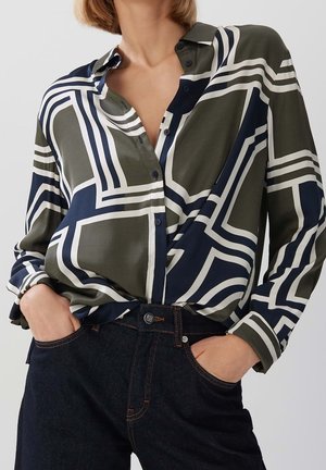 Žena nosící košili s dlouhým rukávem na knoflíky se zelenými, námořnickými a bílými geometrickými vzory, zastrčenou do tmavých džínů z denimu.