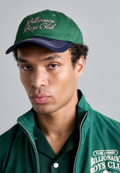 Casquette verte avec visière marine et texte brodé "Billionaire Boys Club." Veste zippée verte avec logo blanc et détails de boutons.