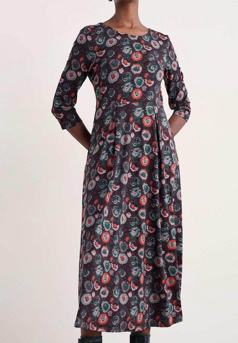 Femme portant une robe à manches longues, longueur mi-longue, avec un motif floral multicolore comprenant des fleurs vertes, rouges et violettes. Mains dans le dos.