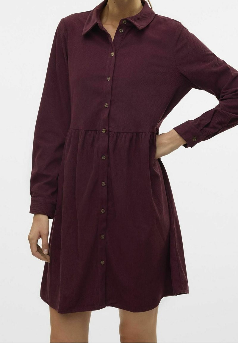 Femme portant une robe en velours côtelé violet foncé à manches longues, boutonnée, avec un col et une couture à la taille, posant une main sur la hanche.