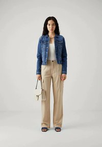 Kvinne med langt mørkt hår, iført en blå denimjakke, hvit topp, beige vide bukser, holder en hvit håndveske og har på seg blå sandaler.