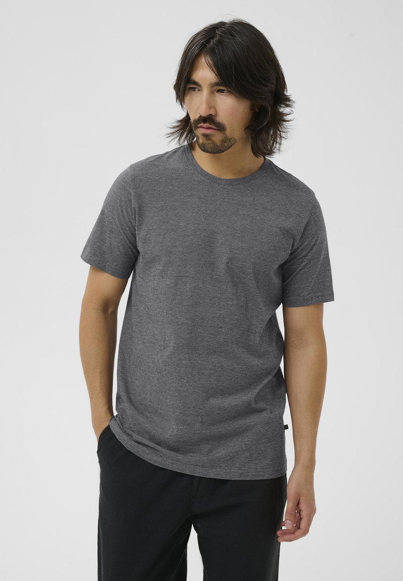 Homme aux cheveux mi-longs et foncés portant un t-shirt gris uni à manches courtes et un pantalon noir, debout devant un fond clair.