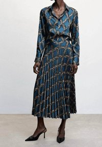 Robe en soie à manches longues, couleur sarcelle, avec un motif répété en beige et noir. Jupe plissée et détail col. Portée avec des escarpins noirs à bouts pointus.