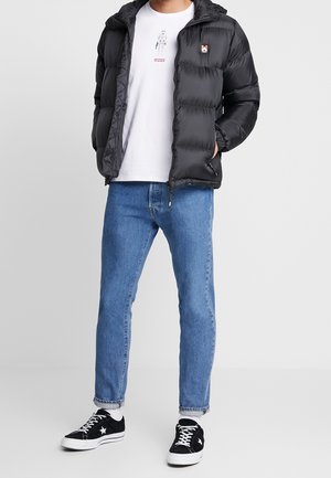 Chaqueta puffer negra con capucha, con cremallera frontal, parche de logo en contraste, corte relajado. Lleva puesta sobre una camiseta gráfica blanca y jeans azules.