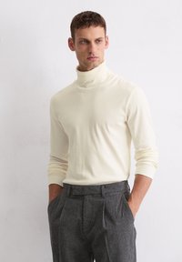 Cremfarbener Rollkragenpullover mit weicher Textur, figurbetontem Design und langen Ärmeln, kombiniert mit grauen Hosen.