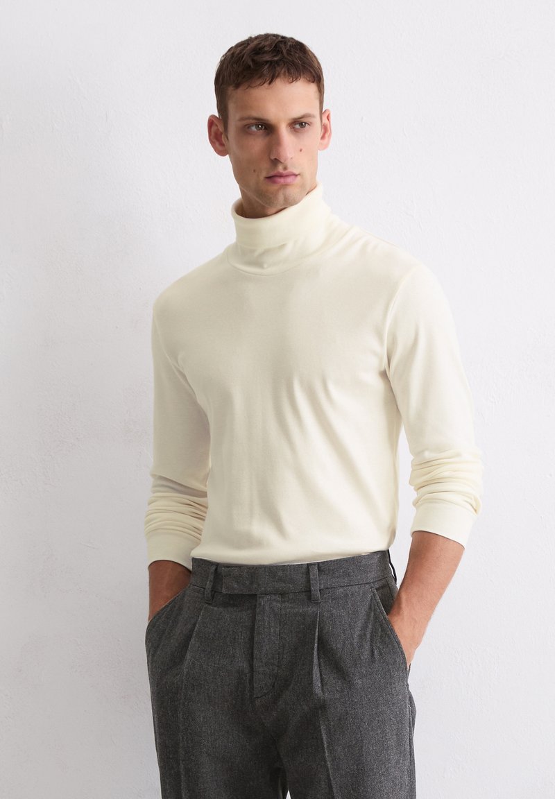 Cremfarbener Rollkragenpullover mit weicher Textur, figurbetontem Design und langen Ärmeln, kombiniert mit grauen Hosen.