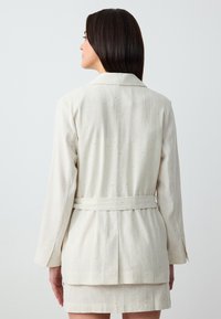Blazer beige chiaro con strisce bianche verticali, tessuto testurizzato, vita con cintura e maniche lunghe. Design sartoriale con colletto classico.