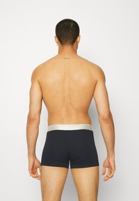 Schwarze Boxershorts mit einem silbernen Bund, von der Rückseite gezeigt. Glattes Material und taillierte Passform, die einen nackten Rücken und Schultern zeigen.