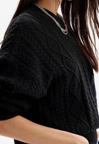 Černý pletený svetr s texturovanými copánkovými vzory, kulatým výstřihem a širokými rukávy. Zvýrazněn vrstveným stříbrným náhrdelníkem.