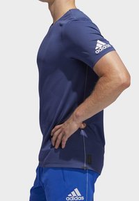 Modrá sportovní košile s texturovanou tkaninou, krátkými rukávy a bílým logem Adidas na levém rukávu. Má kulatý výstřih a tkanou etiketu.