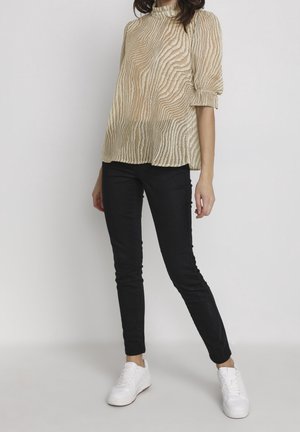 Blouse beige à col montant avec un design texturé en vagues, manches courtes bouffantes, assortie à un jean skinny noir et des baskets blanches.