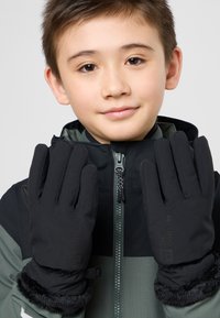 Schwarze Handschuhe mit einer strukturierten Oberfläche und Kunstfellbündchen, die ein Logo am Handgelenk aufweisen. Getragen von einem Kind in einer zipfeljacke.
