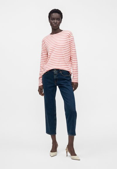 Femme portant une chemise à manches longues rayée rose et blanche, un jean court bleu et des chaussures à talons hauts pointues beiges, debout devant un fond blanc.