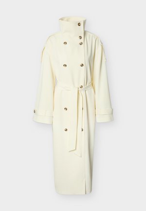 Lang cremefarvet trenchcoat med høj krave, dobbeltradede knapper, bælte i taljen og manchetter med knapper, vist på hvid baggrund.