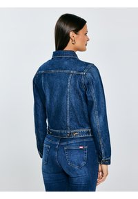 Pikkade tumejuustega naine, kes kannab kuldseid rõngaskõrvarõngaid, tumedat sinist teksajopet ja sobivaid teksapükse, seisab külje peal.