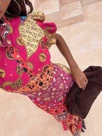 Kleurvolle maxi-jurk met bloemenpatroon en volants mouwen, in roze, oranje en gouden tinten. Vergezeld van een bruine clutch.