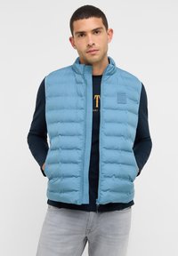 Mann in einer hellblauen Steppweste über einem navyblauen Langarmshirt und hellgrauen Jeans, stehend mit den Händen in den Weste-Taschen.