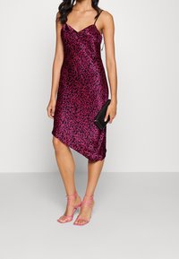 Fuksjowa satynowa sukienka z falistym czarnym wzorem, asymetrycznym dołem i cienkimi ramiączkami, zestawiona z różowymi sandałami na paski i czarną kopertówką.