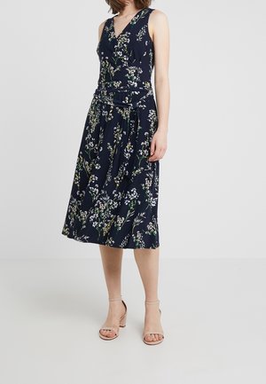 Robe bleu marine sans manches avec un motif floral, design portefeuille, et ceinture à la taille. Jupe fluide tombant sous le genou. Associée à des talons de couleur claire.