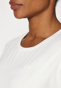 Camiseta blanca de manga corta con escote redondeado, que presenta un logo sutil en gris claro en el pecho. Tela lisa y ligera.