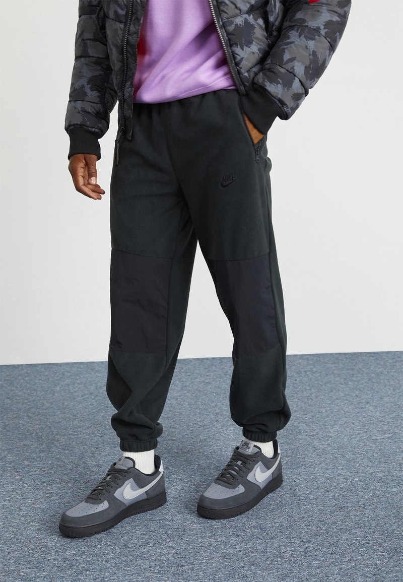 Pantaloni in fleece neri e grigio scuro con una finitura testurizzata, dotati di polsini affusolati e un piccolo logo Nike sulla parte anteriore. Abbinati a sneaker grigie.