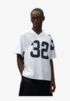 Persona che indossa una maglia sportiva bianca in rete con i numeri "32" in blu scuro e pantaloncini neri, in piedi con le mani parzialmente nelle tasche.