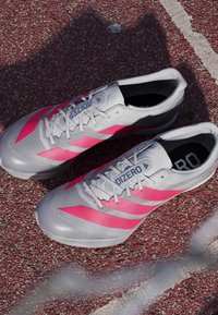 Harmaat juoksukengät, joissa on vaaleanpunaisia yksityiskohtia, teksturoitu materiaali, erottuvat nauhat ja näkyvä brändäys sivussa, asetettuna punaiselle pinnalle.