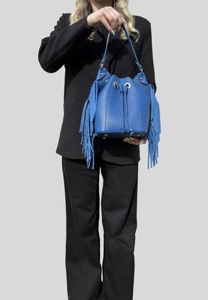 Persona con abbigliamento nero tiene una borsa a secchiello in pelle blu con dettagli a frange e borchie di metallo sul fondo, su sfondo grigio.