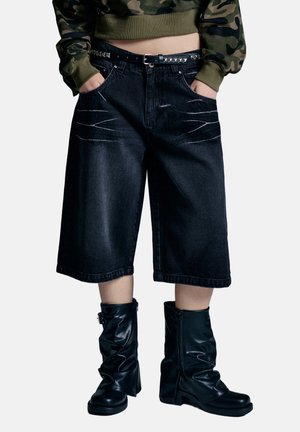 Person wearing black wide-leg denim shorts with white stitching, a studded black belt, camouflage cropped sweatshirt, and black leather boots.