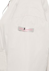 Manica di giacca beige chiaro con un piccolo etichetta grigia che mostra un'icona di cane nero e un testo rosa che dice "Frieda & Freddies New York."