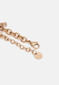 Bracelet en chaîne dorée rose avec des maillons entrelacés, un fermoir en forme de homard et une étiquette ronde arborant un logo. Finition lisse avec un éclat subtil.