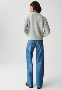 Frau steht mit dem Rücken zur Kamera, trägt einen grauen Rippenpullover, blaue weite Jeans und schwarze Schuhe vor einem schlichten hellen Hintergrund.