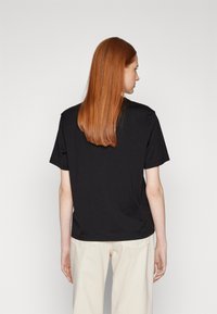 Schwarzes Baumwoll-T-Shirt mit kurzen Ärmeln, lockerer Passform und rundem Ausschnitt, von hinten betrachtet. Wird mit hellen Hosen kombiniert.
