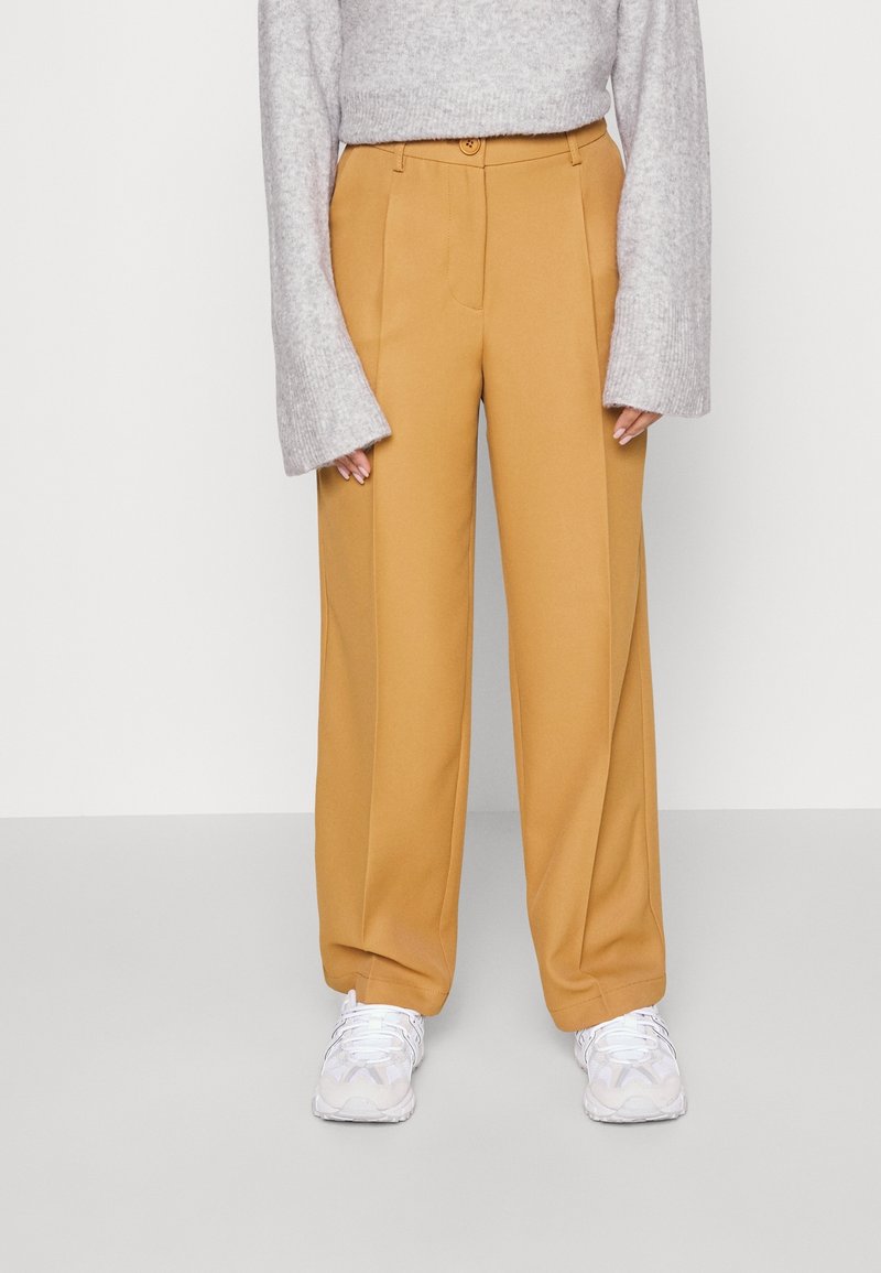 Femme portant un pantalon large moutarde, un pull gris à manches longues avec des poignets évasés, et des baskets blanches, debout devant un fond uni.