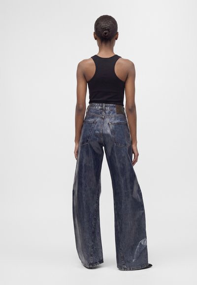 Femme avec les cheveux en chignon portant un haut noir sans manches et un jean bleu délavé à jambes larges, vue de dos sur un fond uni.