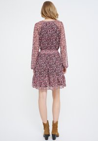 Florale chiffon jurk met lange mouwen, gatherings in de taille en een gelaagde rok in tinten roze en paars. Bruine haklaarzen complementeren de outfit.