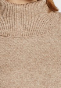 Beżowy sweter z golfem o ryżowym splocie. Miękka, dziana tkanina z podwiniętym, wysokim kołnierzem i gładkim wykończeniem.