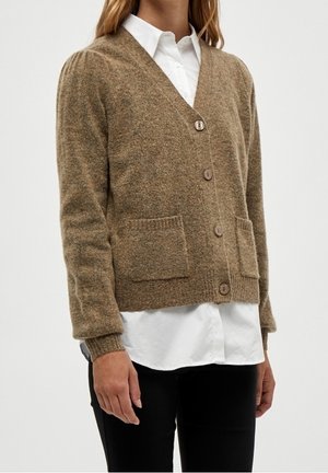 Femme portant un cardigan marron boutonné avec deux poches avant, par-dessus un chemisier blanc et un pantalon noir, debout devant un fond uni.