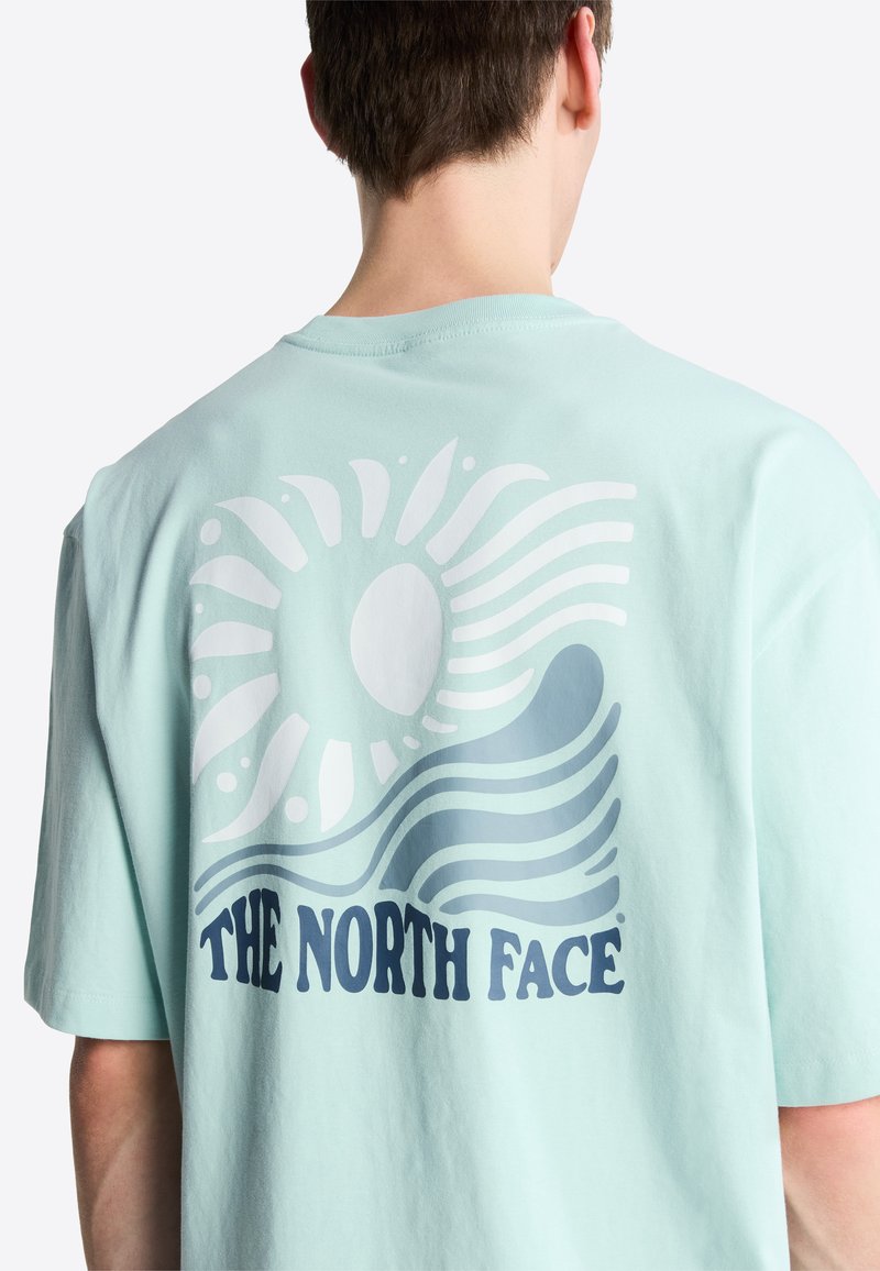 Person trägt ein hellblaues T-Shirt mit weißem und dunkelblauem Sonnen- und Wellenmotiv sowie dem Schriftzug "THE NORTH FACE" auf der Rückseite.