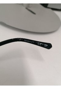 Montura de gafas negra con una patilla curva. Presenta texto blanco con el nombre de la marca en el lateral, sobre un acabado mate.