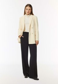 Helle beige Corduroy-Blazer mit Doppelknopf, kombiniert mit einem cremefarbenen Rollkragenoberteil und schwarzen weit geschnittenen Hosen. Schwarze High-Heels.